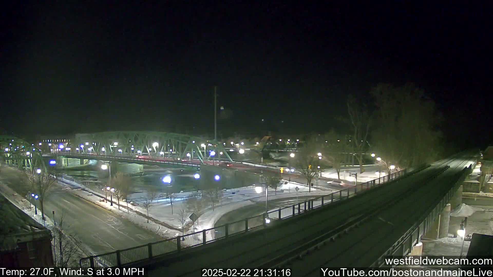 A nighttime view of a snow-covered city scene featuring a bridge over a river, with a few cars on the roads and a train track in the foreground, under a clear, dark sky with a south wind at 3 mph and a temperature of 27 degrees Fahrenheit.