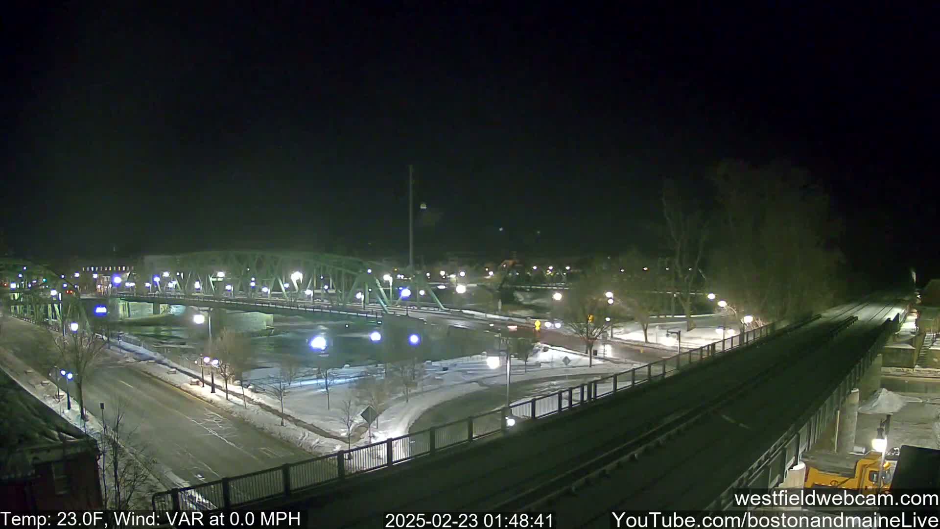 A snow-covered nighttime scene shows a bridge crossing a river, with roads and streetlights illuminated under a clear, dark sky.