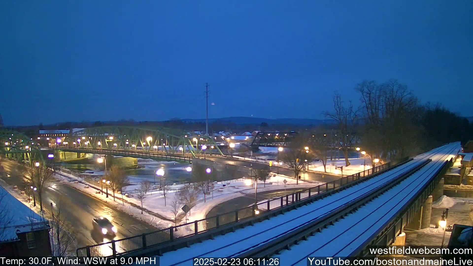 A snow-covered town at twilight features a lit green bridge spanning a river, with train tracks visible in the foreground under a dark blue sky.