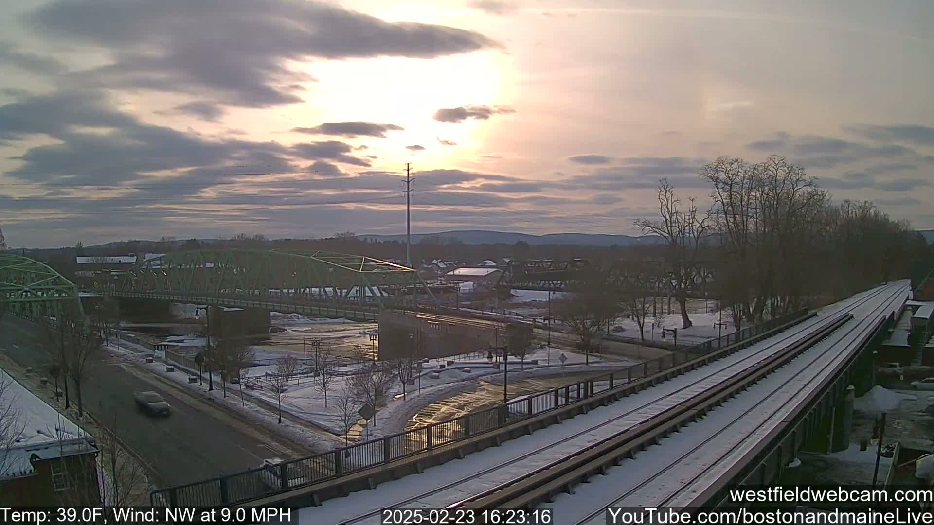 A partly cloudy sunset illuminates a snow-covered town scene with a river, two bridges, and train tracks.