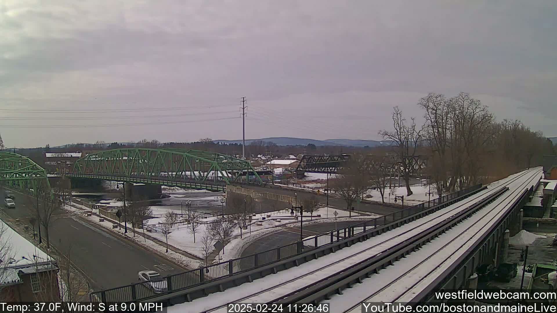 A snow-covered train track stretches towards two green bridges spanning a river, with a town and distant hills in the background under a cloudy sky.