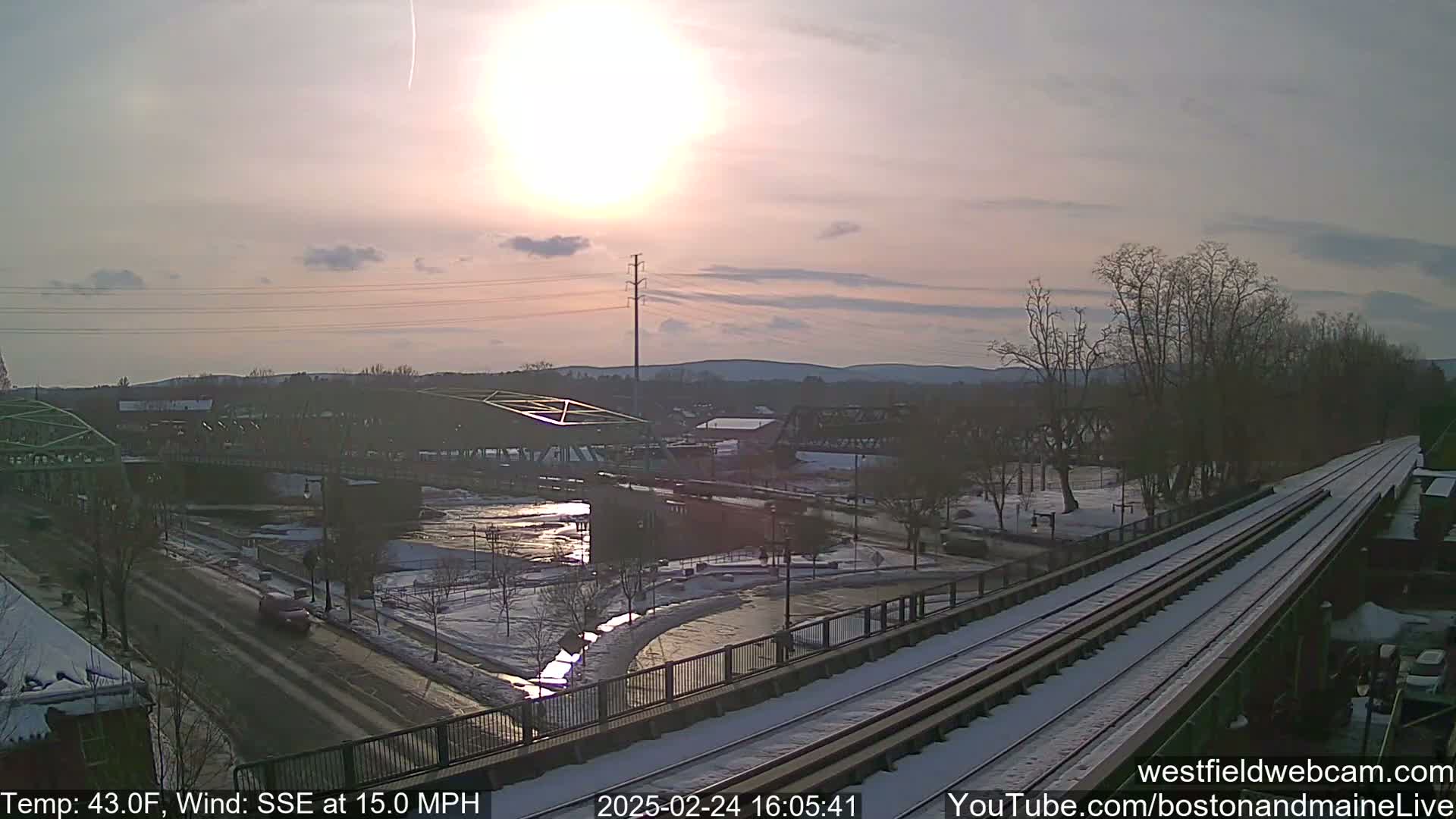 A partly sunny winter scene shows a river partially frozen over, bridges, train tracks partially covered in snow, and a road with cars driving on it.