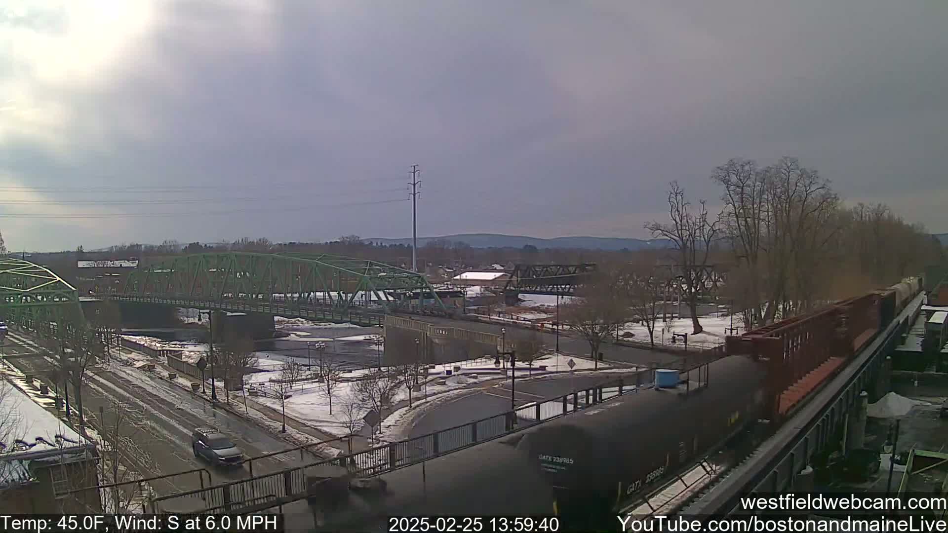 A partly cloudy day with a south wind shows a view of a town with two bridges over a river, one green and one dark metal, with a train passing by in the foreground, and some snow on the ground.