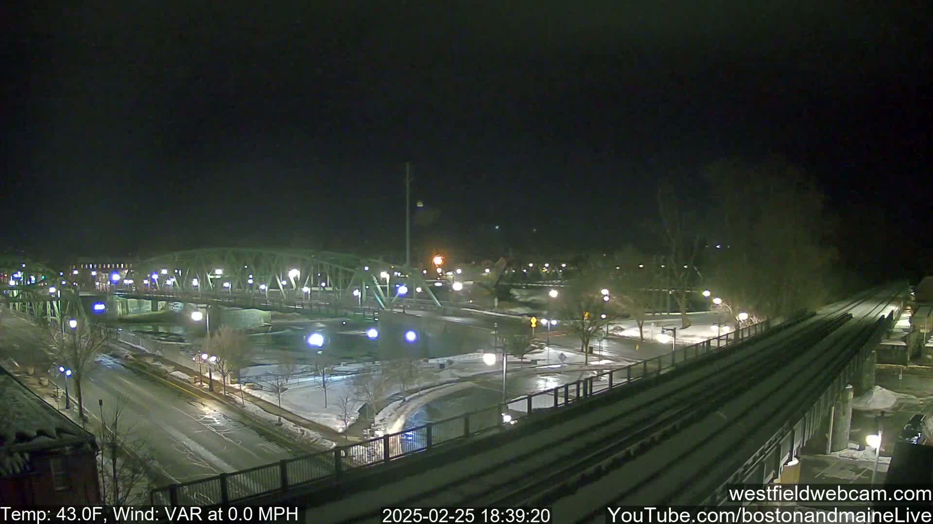 A nighttime view of a bridge over a river, with roads and train tracks, partially covered in snow, under a dark sky.
