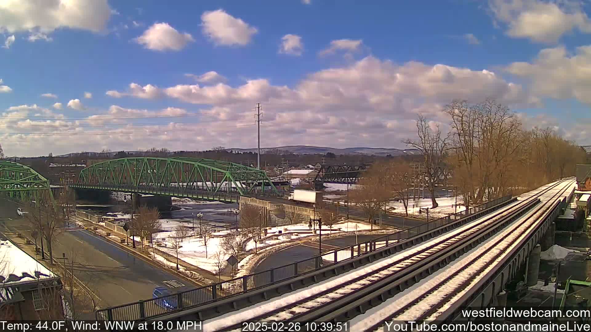 A partly cloudy day with a brisk wind shows a view of a river, two bridges, a roadway, and train tracks partially covered in snow.