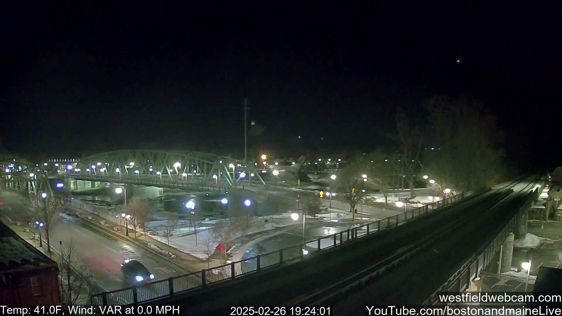 A nighttime view of a bridge over a river, with some cars on roads and light snow on the ground under a clear sky.