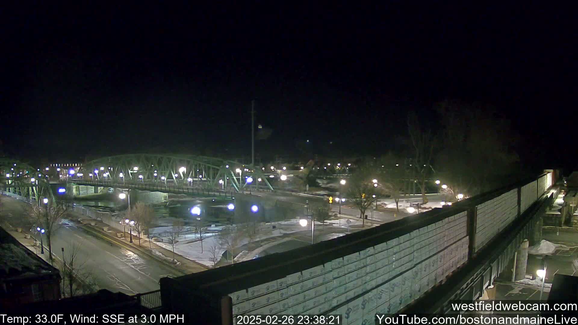 A nighttime view of a lit-up bridge over a river, with a road and sparsely-populated park-like area visible, under a clear, dark sky and a 33°F temperature with a 3.0 mph SSE wind.