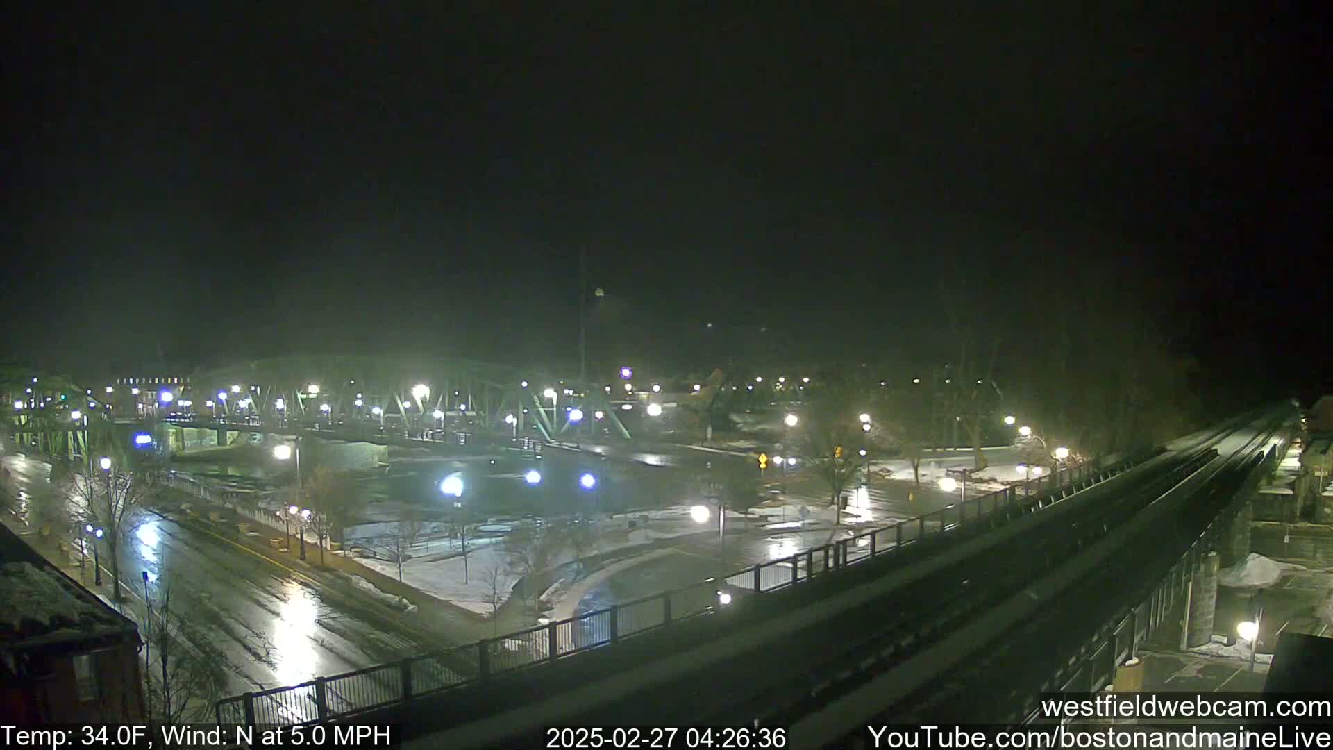 A nighttime view of a bridge and roadway with patches of snow and ice under a dark sky, with a light north wind.