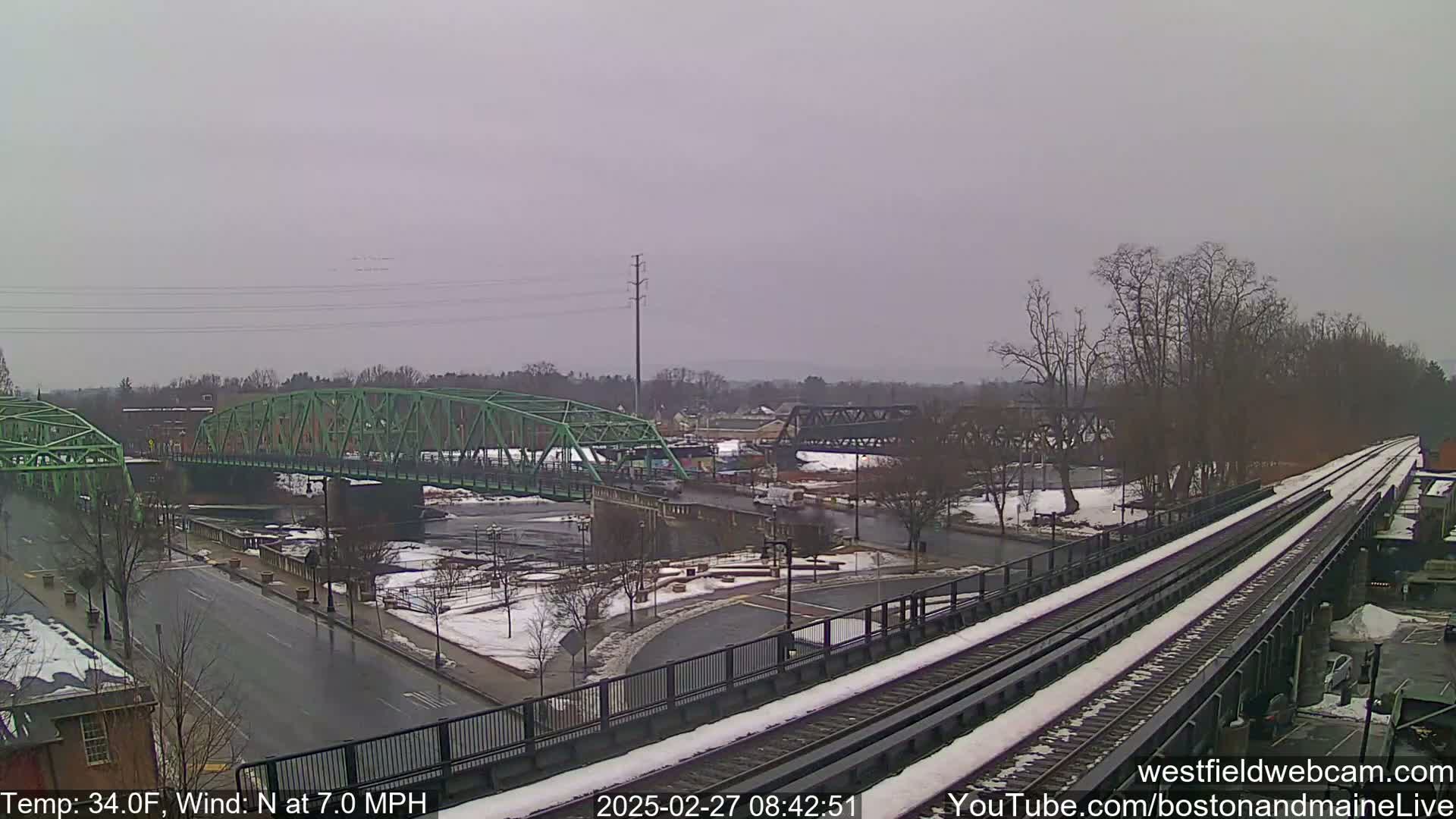 A partly snow-covered town scene with two bridges over a river, viewed from a train trestle on an overcast day.