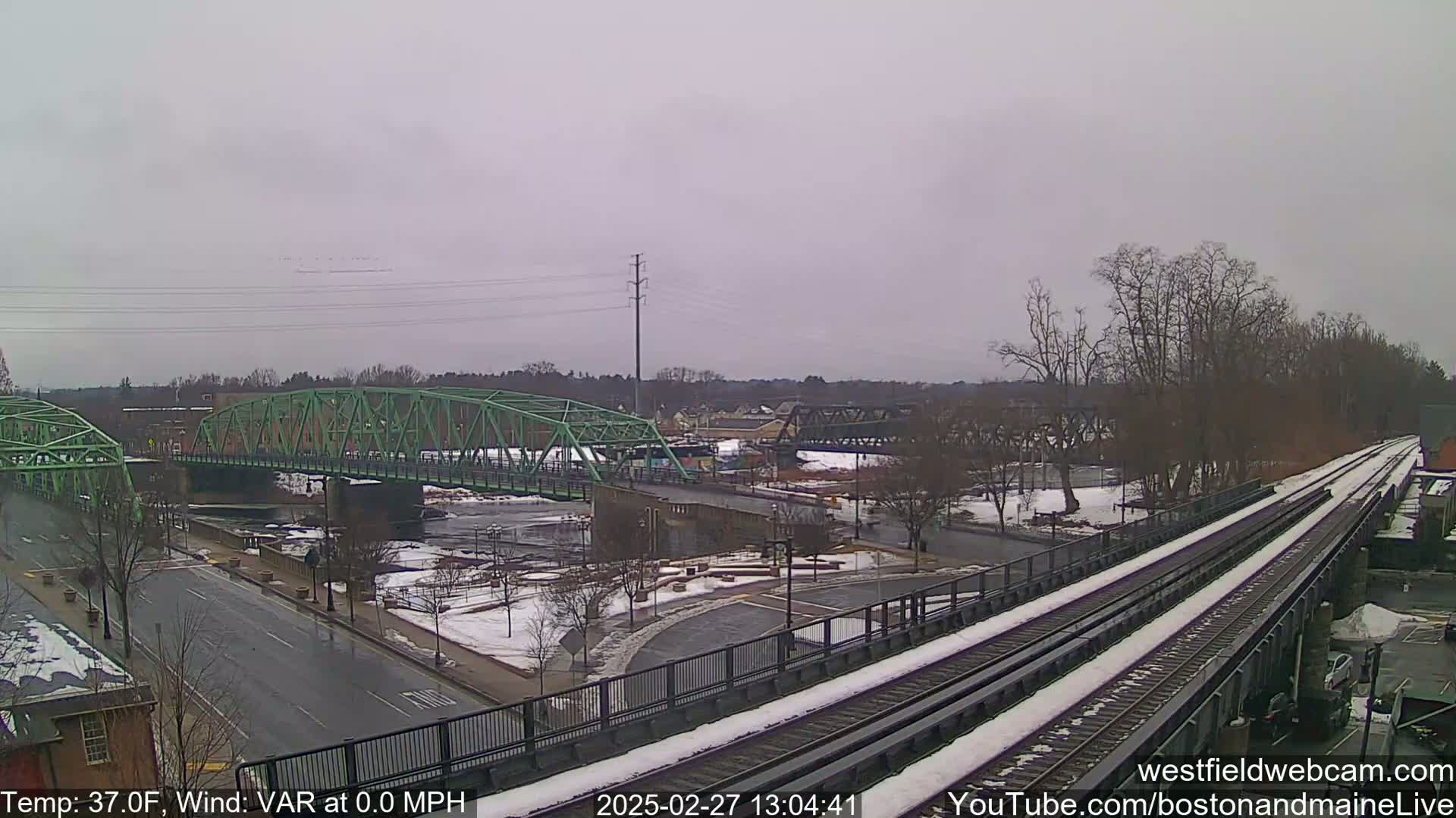A green bridge spans a river partially covered in ice, with a train track running alongside a snow-covered roadway under an overcast sky.