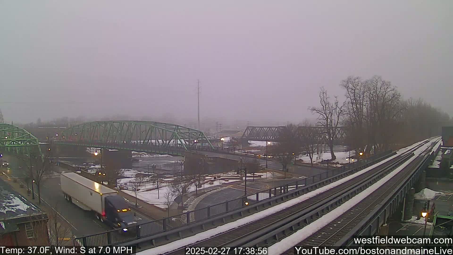 A snow-covered train track runs alongside a river, featuring two bridges and a partially snow-covered roadway under an overcast sky.