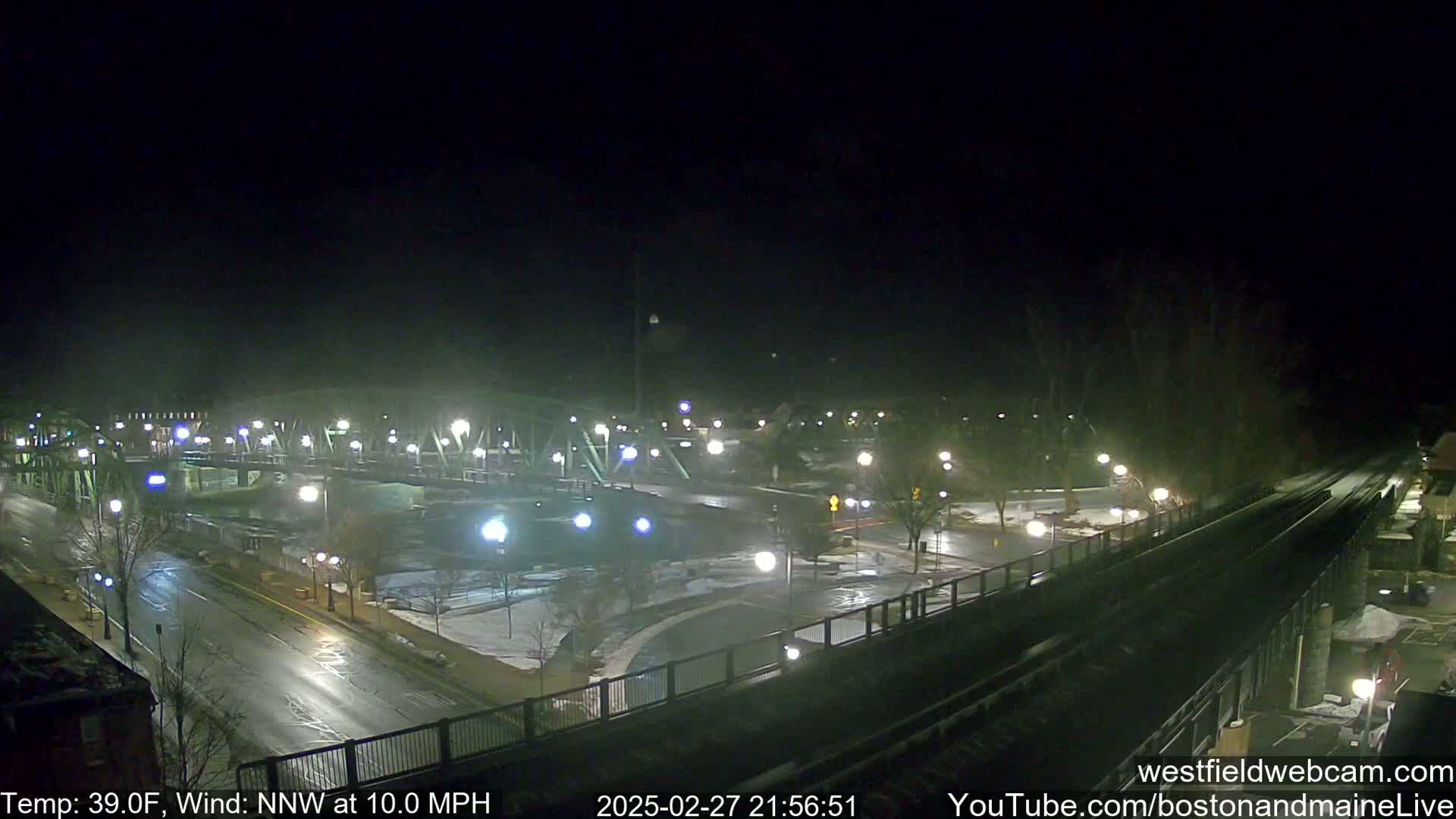 A nighttime view of a bridge and roadway with some snow and ice on the ground, under a dark, clear sky with a north-northwest wind at 10 mph and a temperature of 39°F.