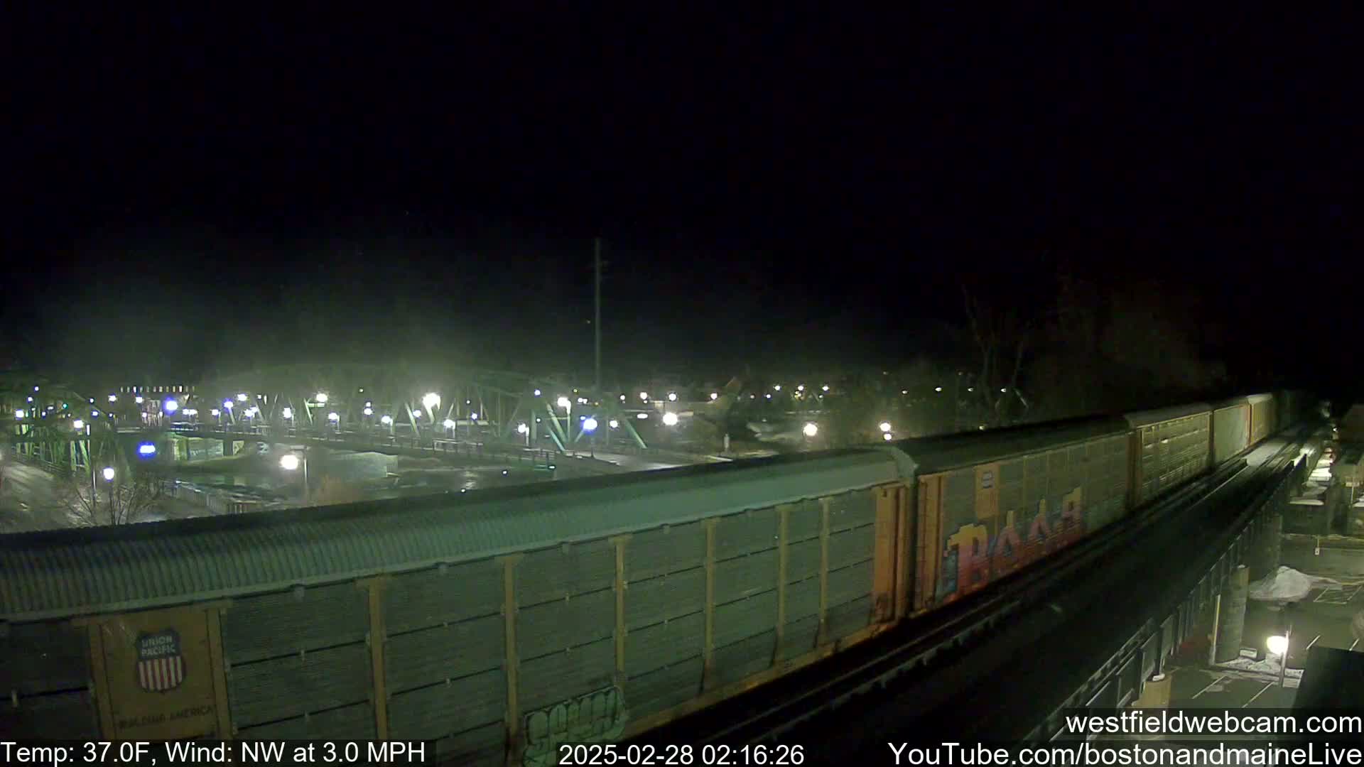 A long freight train passes under a lit bridge at night in 37°F weather with a northwest wind of 3.0 mph.