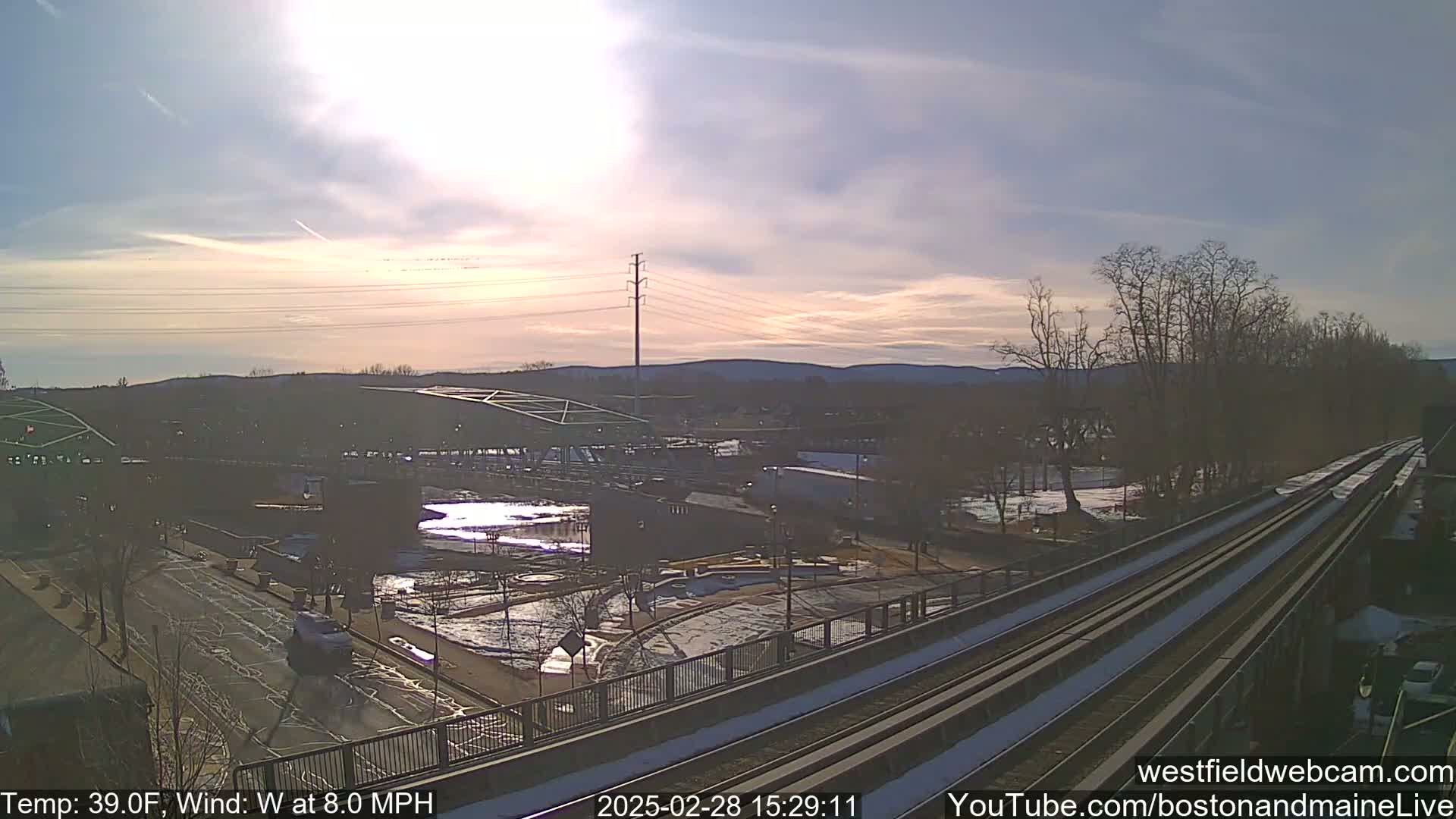 A partly sunny view of a town with a river, train tracks, and a bridge, under a mostly clear sky with a light wind.