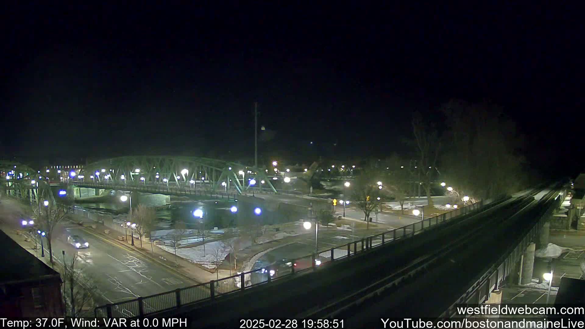 A nighttime view of a bridge over a river, with a roadway and train tracks visible, under clear skies and a temperature of 37°F.