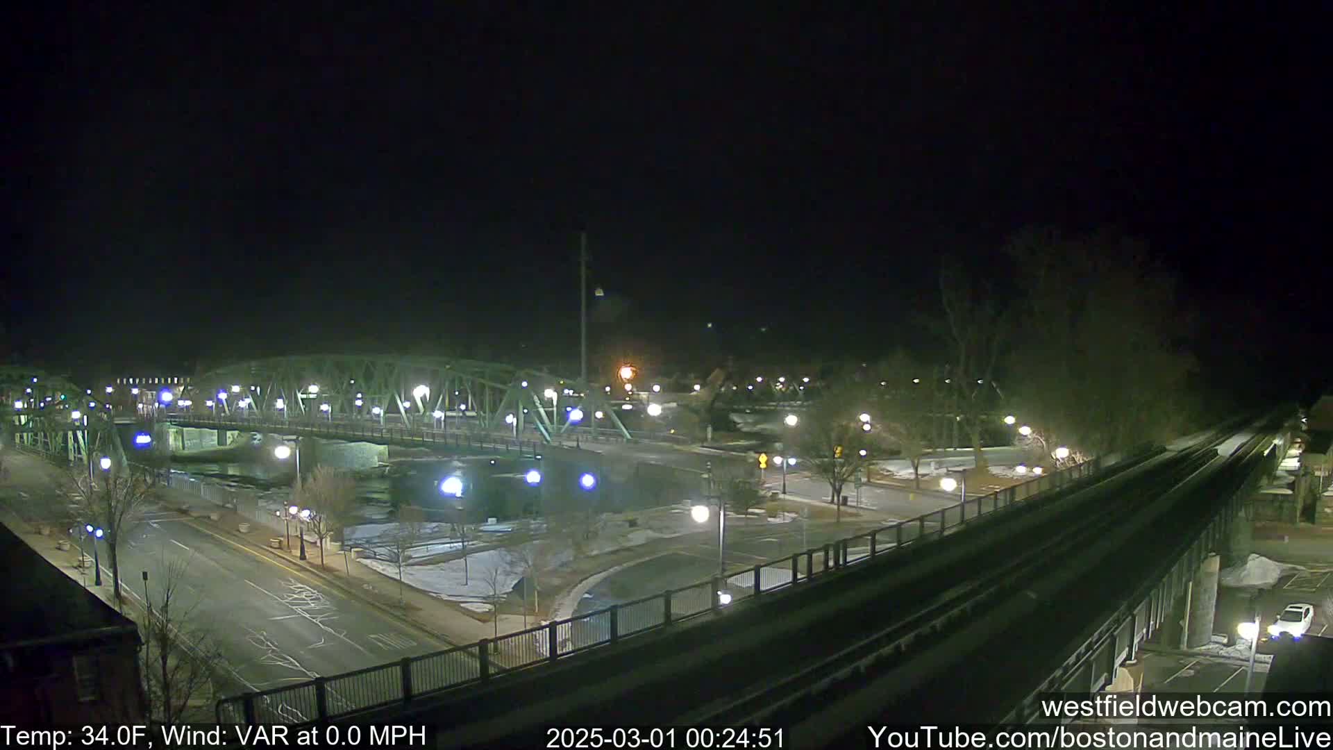 A nighttime view of a bridge over a river, with a road and train tracks visible, under clear skies and a temperature of 34 degrees Fahrenheit.