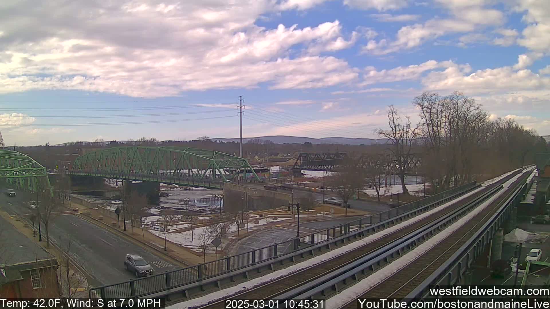 A view of two bridges over a river, with a train track in the foreground, under partly cloudy skies and a temperature of 42°F, with a south wind at 7 mph.