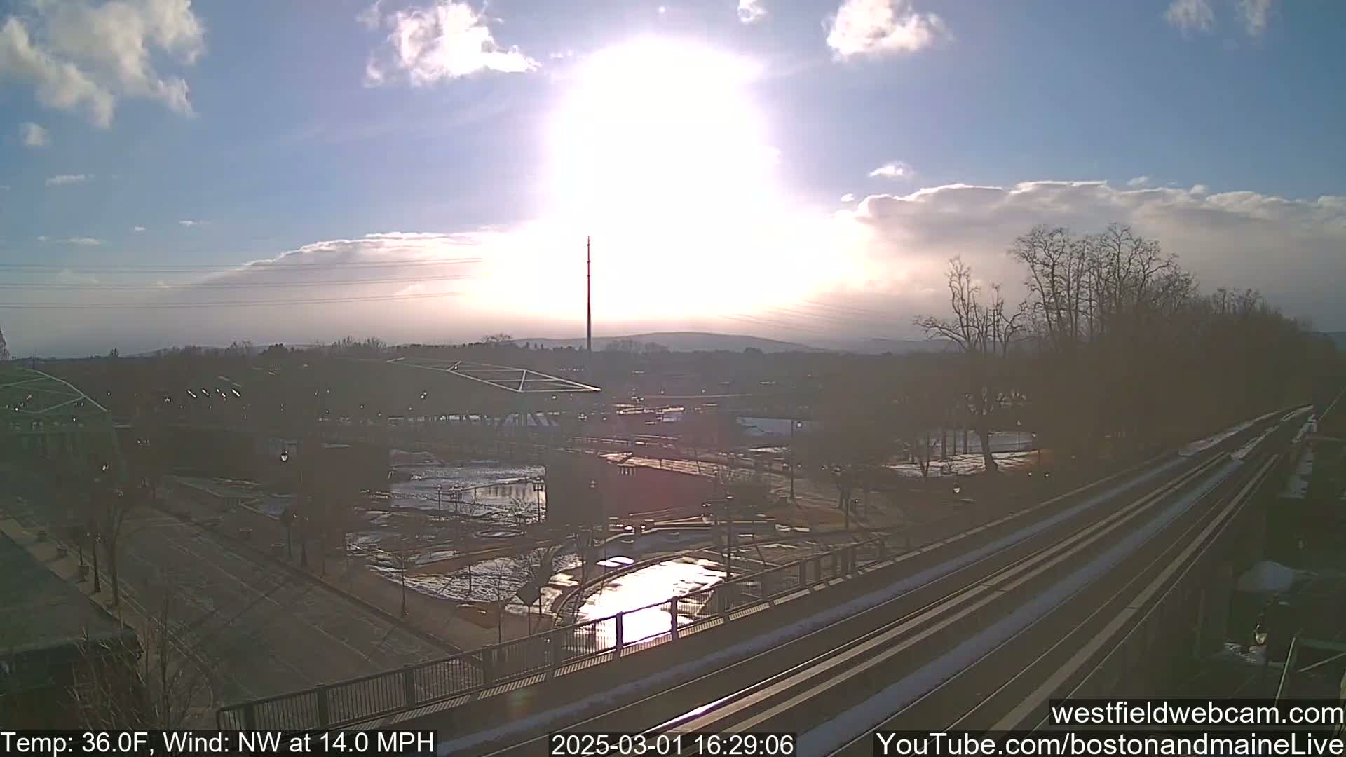 A partly sunny, cold day with a northwest wind shows a view of a train track and urban landscape with patches of snow and ice visible on the ground.
