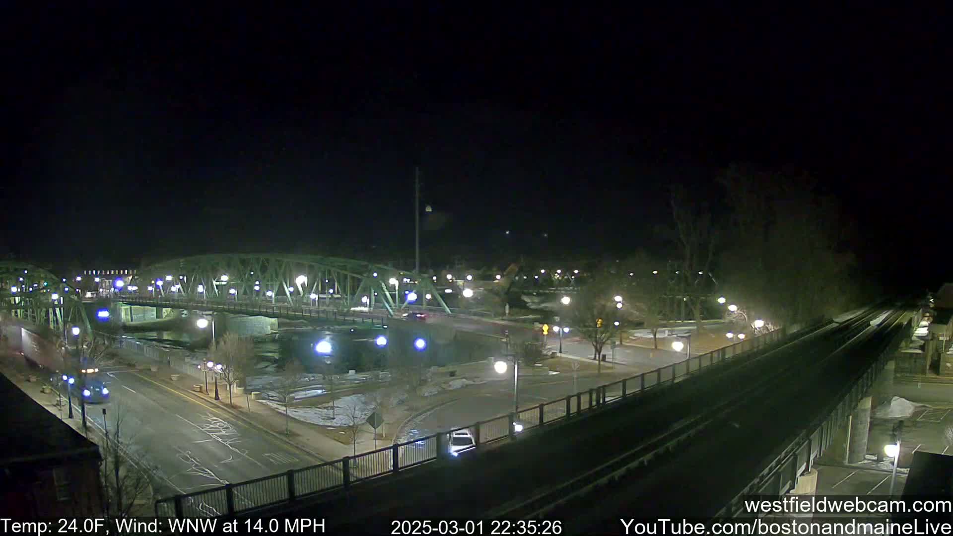 A nighttime view of a bridge over a river, with a road and train tracks intersecting near a park, under a dark, clear sky with a temperature of 24°F and a wind of 14 mph from the west-northwest.