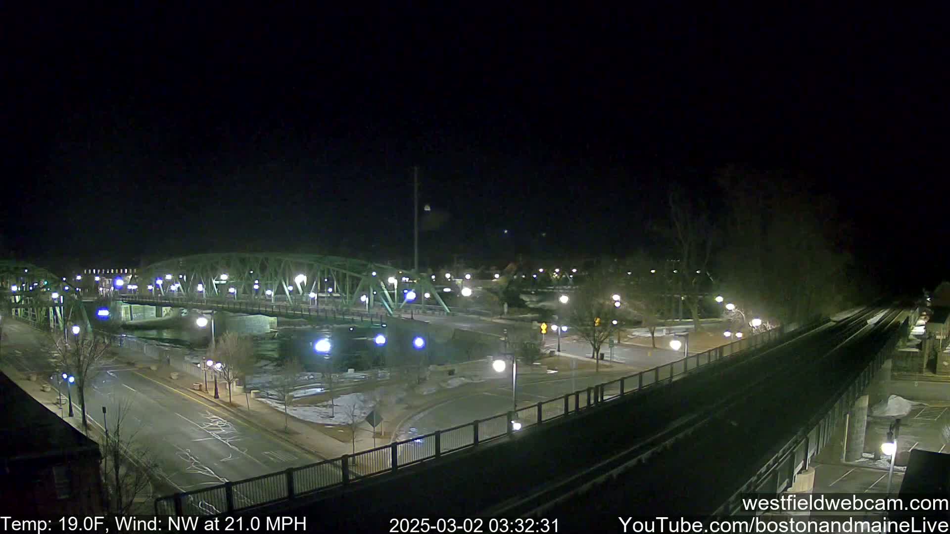 A nighttime view of a bridge over a river, with a train track and roadway intersecting nearby, under a dark, clear sky with a strong northwest wind.