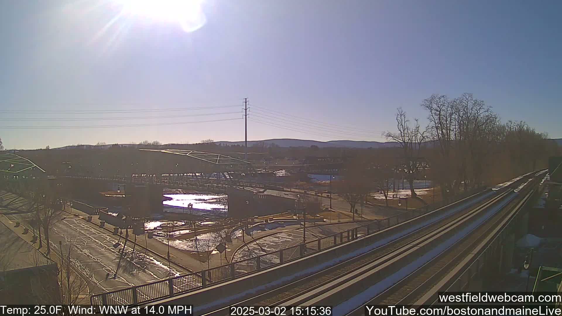 A sunny, cold day with a WNW wind shows a view of a bridge, a roadway, and train tracks, with some snow and ice visible on the ground.