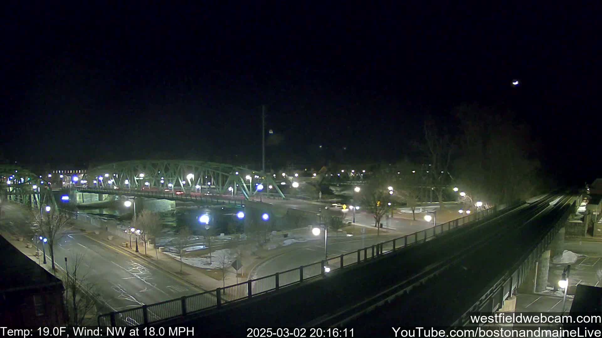 A nighttime view of a bridge over a river, with a road and train tracks visible, under a dark sky with a northwest wind blowing at 18 mph and a temperature of 19°F.