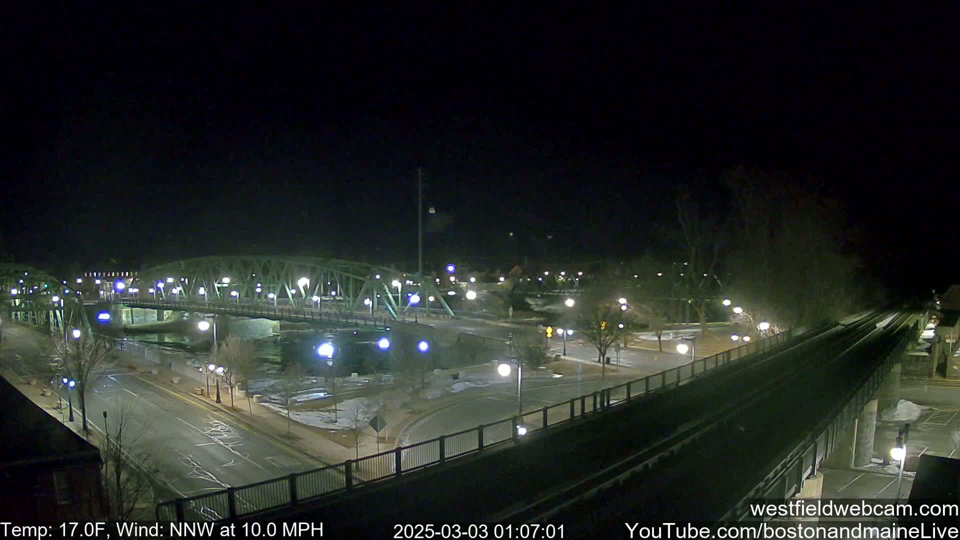 A nighttime view of a bridge over a river, with a road and train tracks intersecting, under a clear, cold, windy sky.