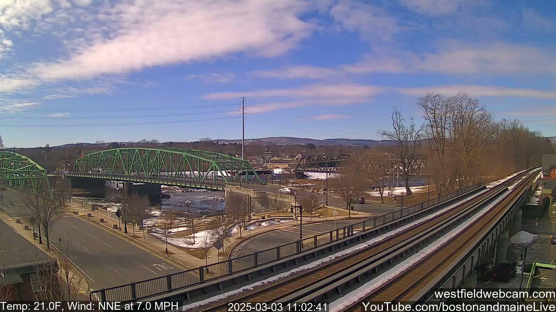 A green bridge spans a river, with a train track and road alongside, under a partly sunny sky with a light wind.