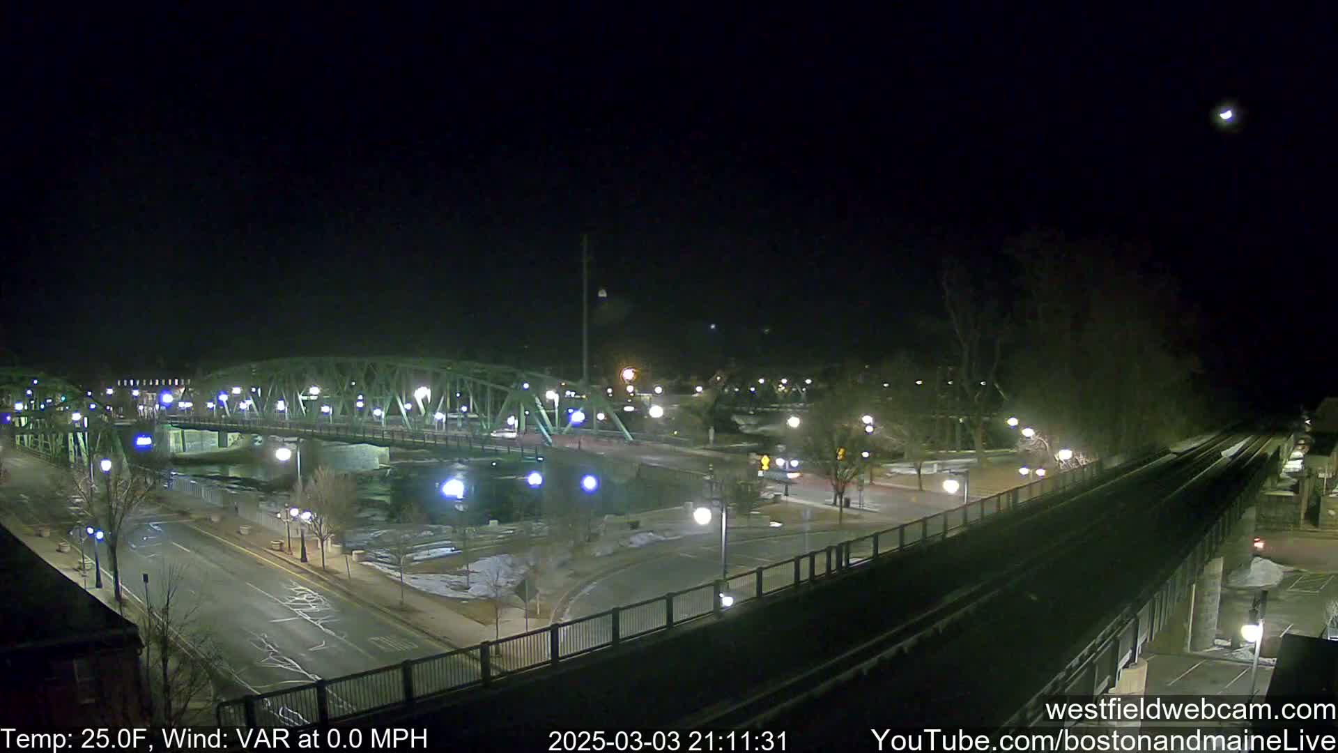 A nighttime view of a bridge over a river, with a road and train tracks visible, under a clear sky.