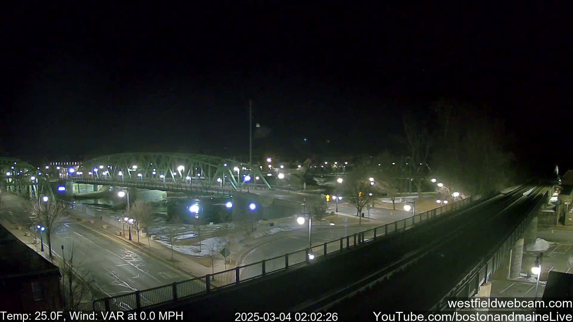 A nighttime view of a bridge over a river, with a road and train tracks visible, under clear skies and a temperature of 25 degrees Fahrenheit.