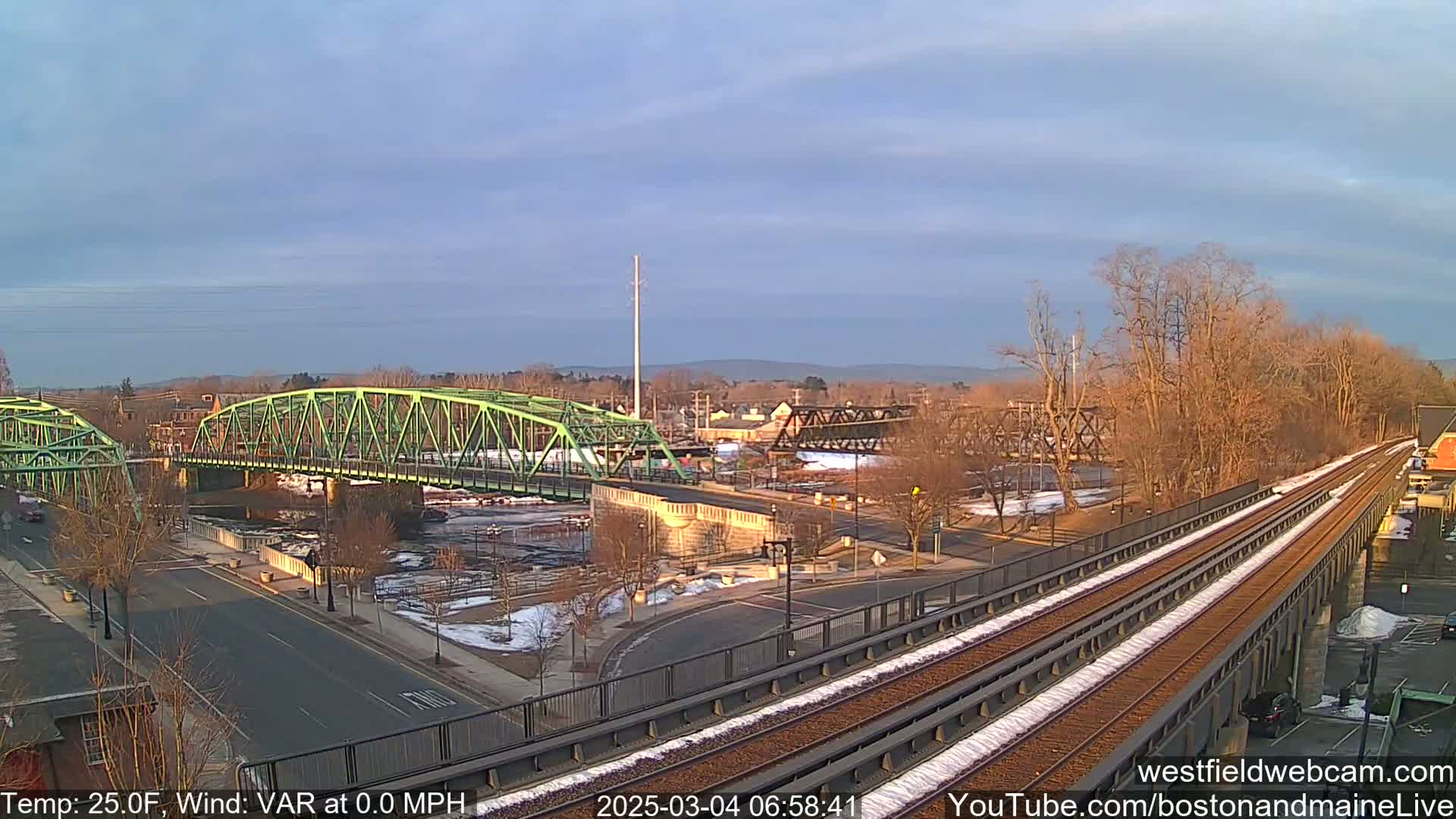 A high-angle view shows two bridges over a river, one green and one dark metal, with train tracks running alongside, a road crossing the scene, and some snow visible on the ground under a clear, bright sky.