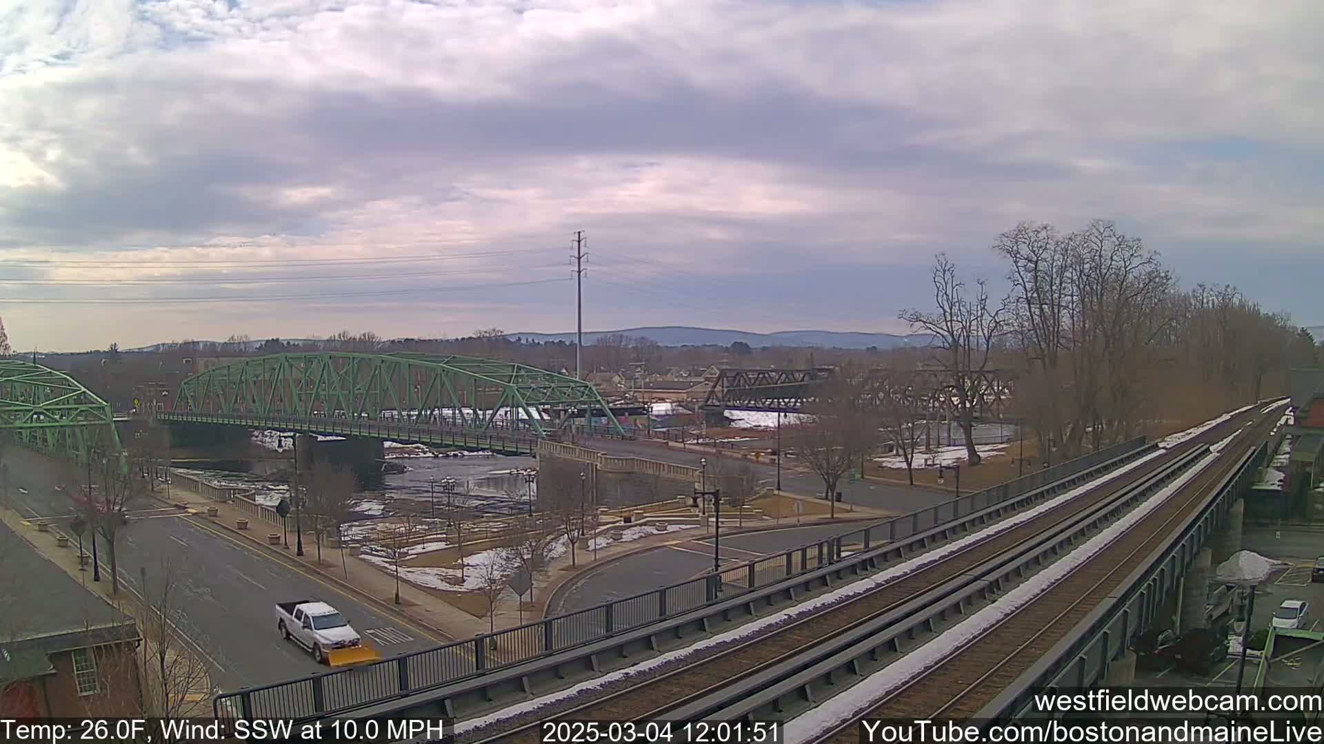 A view of a river with two bridges, one green and one dark metal, under a cloudy sky with a light dusting of snow on the ground and train tracks in the foreground.