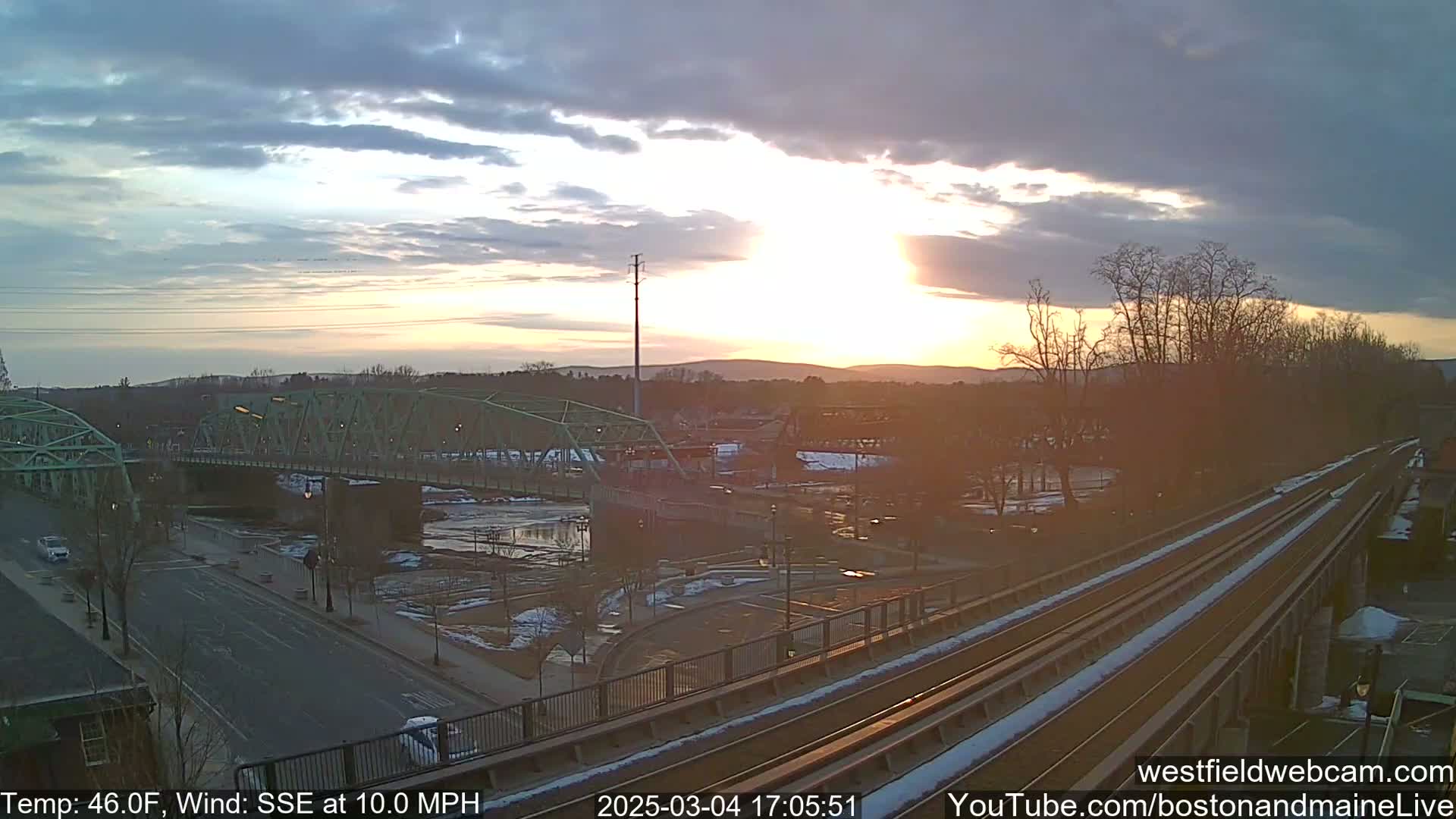 A partly cloudy sunset illuminates a green bridge over a river, with a train track and road visible in the foreground, and some patches of snow or ice on the ground.