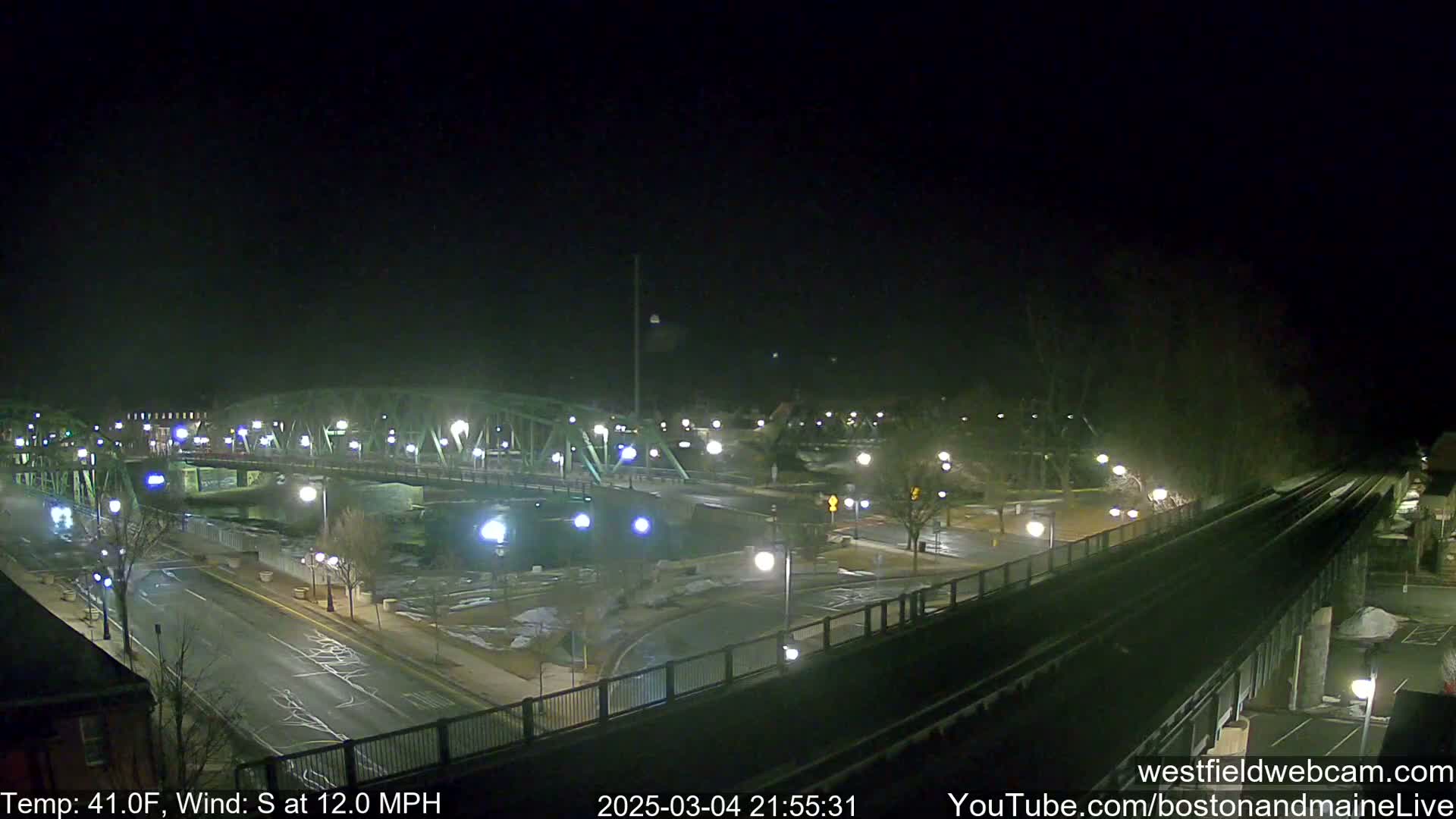 A nighttime view of a bridge over a river, with roads and streetlights illuminated under a dark sky and a south wind at 12 mph.