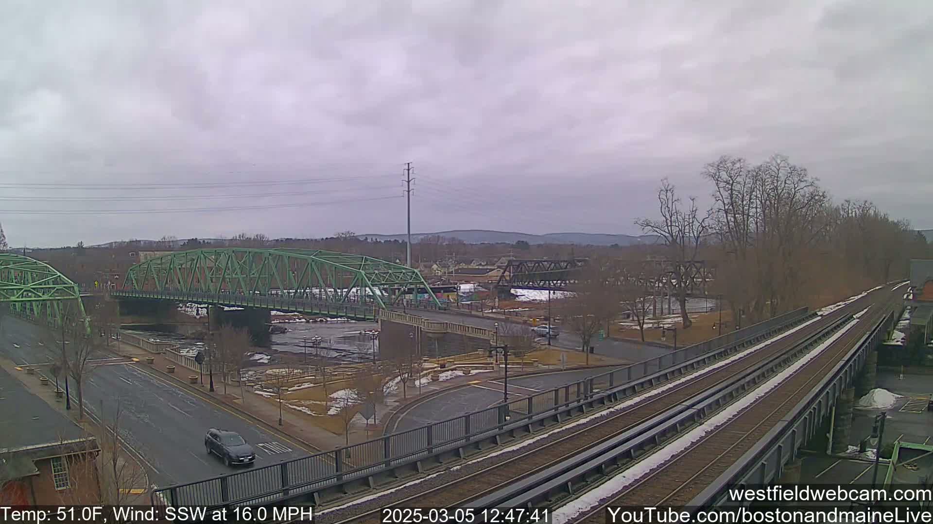 A view of two bridges over a river, one green and one dark metal, with a train track and a road in the foreground on an overcast day with a 16 mph wind.