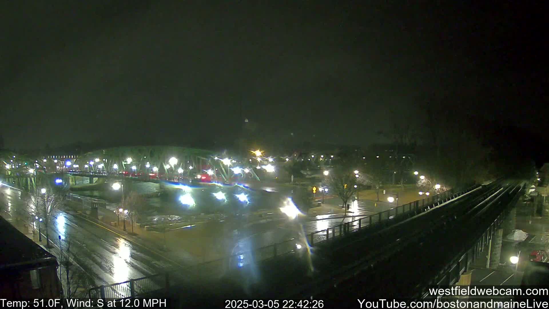 A nighttime view of a rain-slicked city street and bridge, with cars driving across the bridge and streetlights illuminating the wet pavement under a dark sky with a south wind.