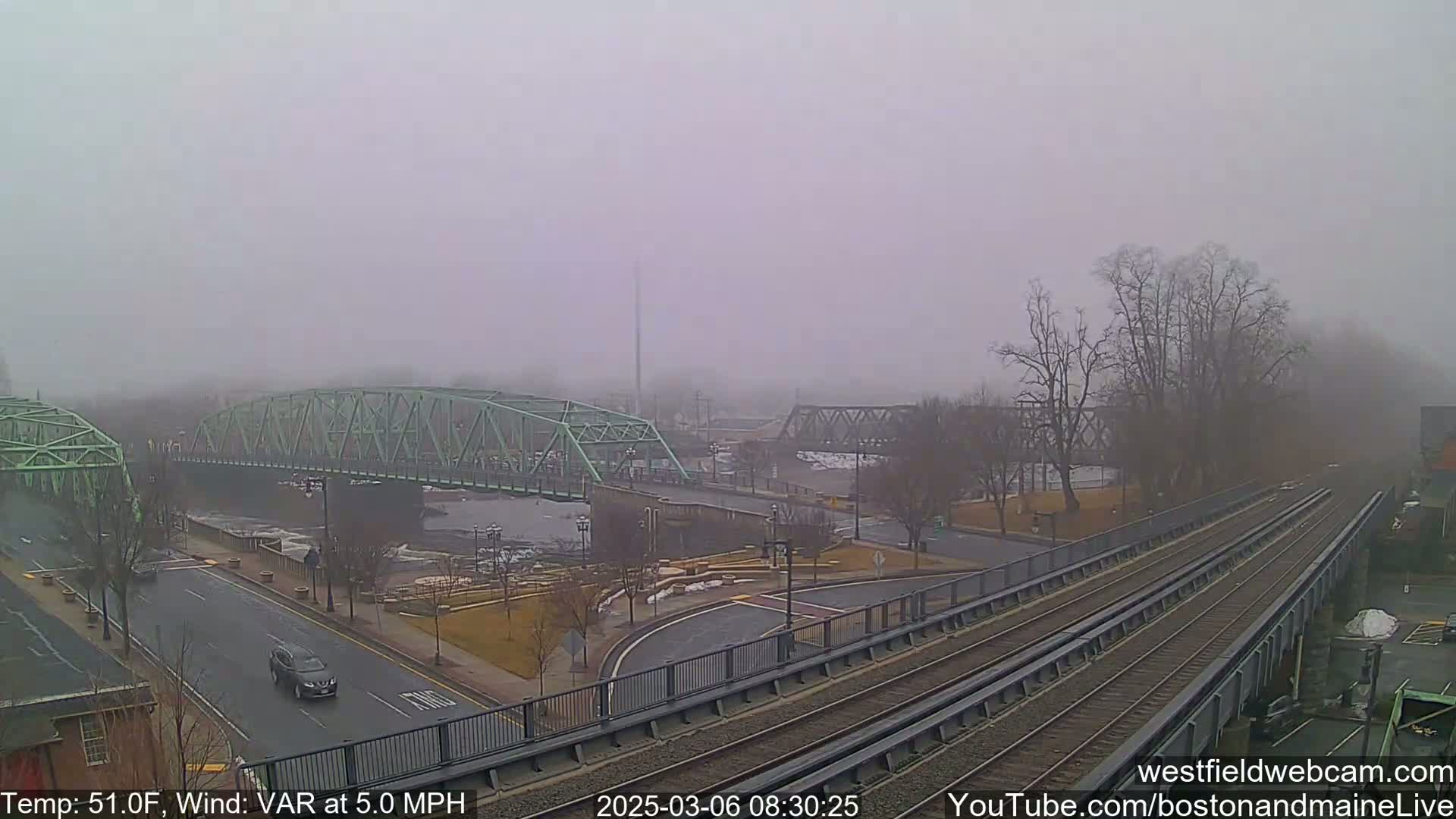 A foggy scene shows two bridges over a river, a roadway with sparse traffic, and train tracks in the foreground.
