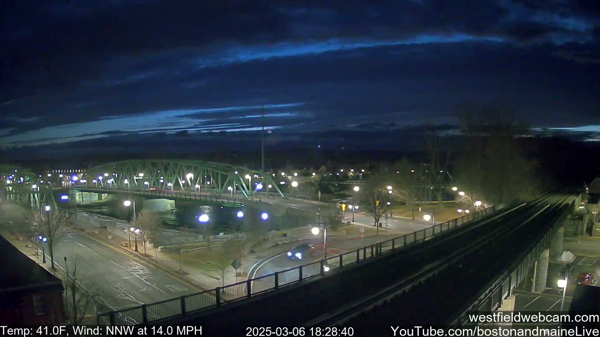 A nighttime view of a bridge over a river, with a road and train tracks below, under a dark blue sky with some clouds and a 14 mph northwest wind.