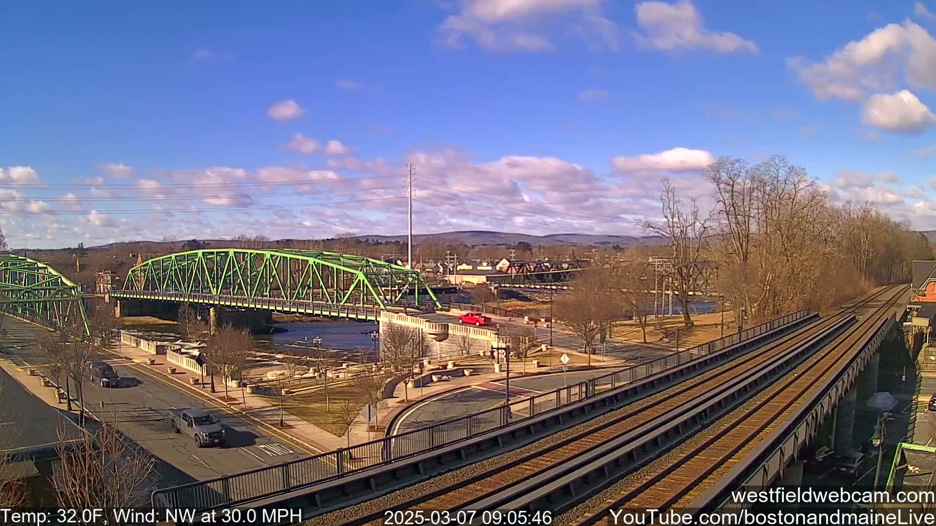 A green bridge spans a river in a town under a partly cloudy sky with a 30 mph northwest wind and a temperature of 32 degrees Fahrenheit; railroad tracks run parallel to the river.