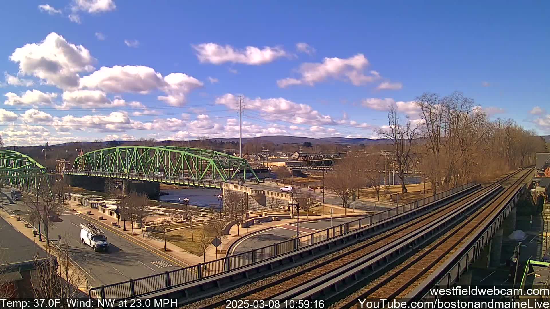 A green bridge spans a river in a town under a partly cloudy sky, with a train track running alongside.