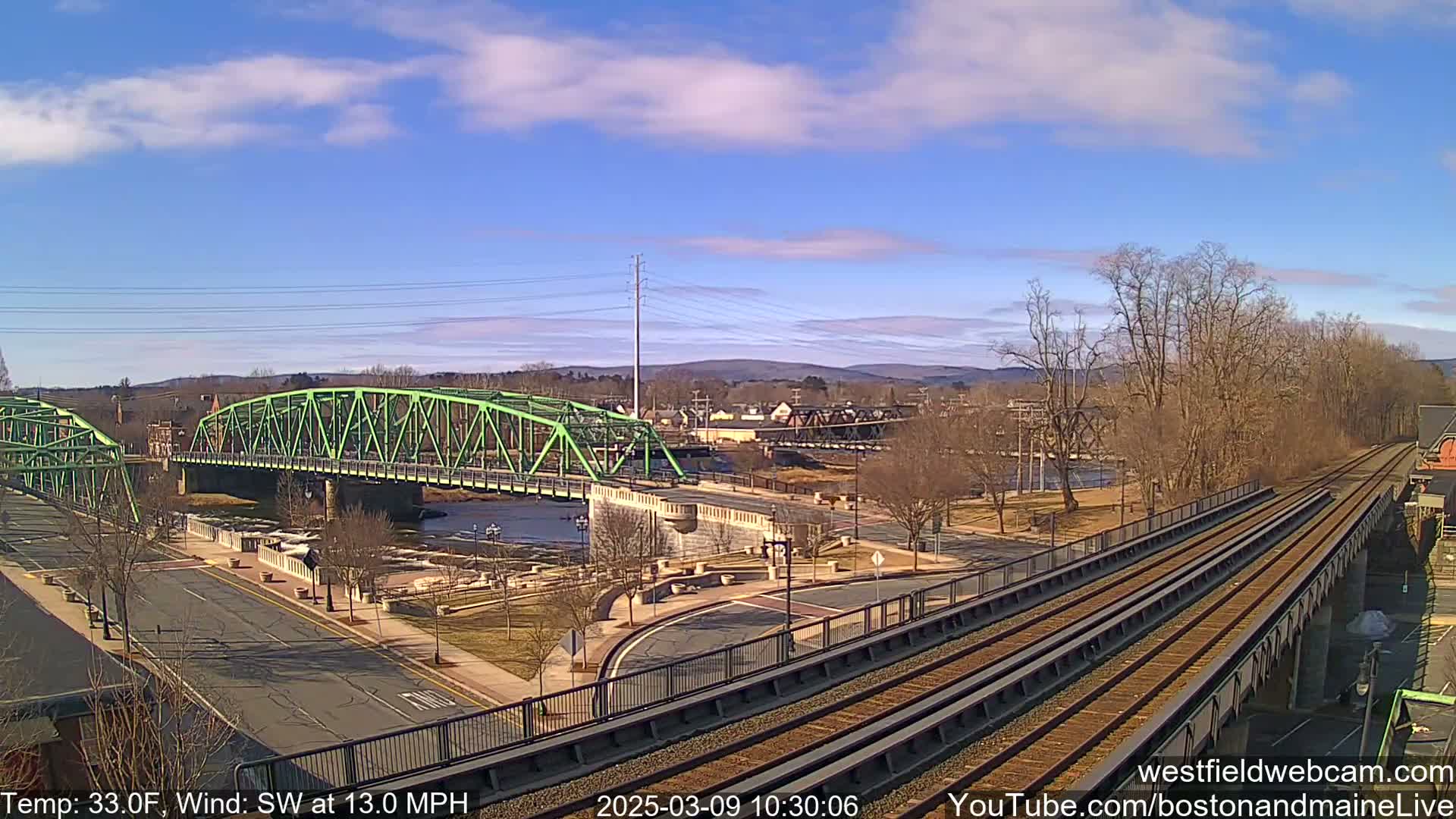 A green bridge spans a river, with train tracks and a roadway leading to it under a partly cloudy sky.