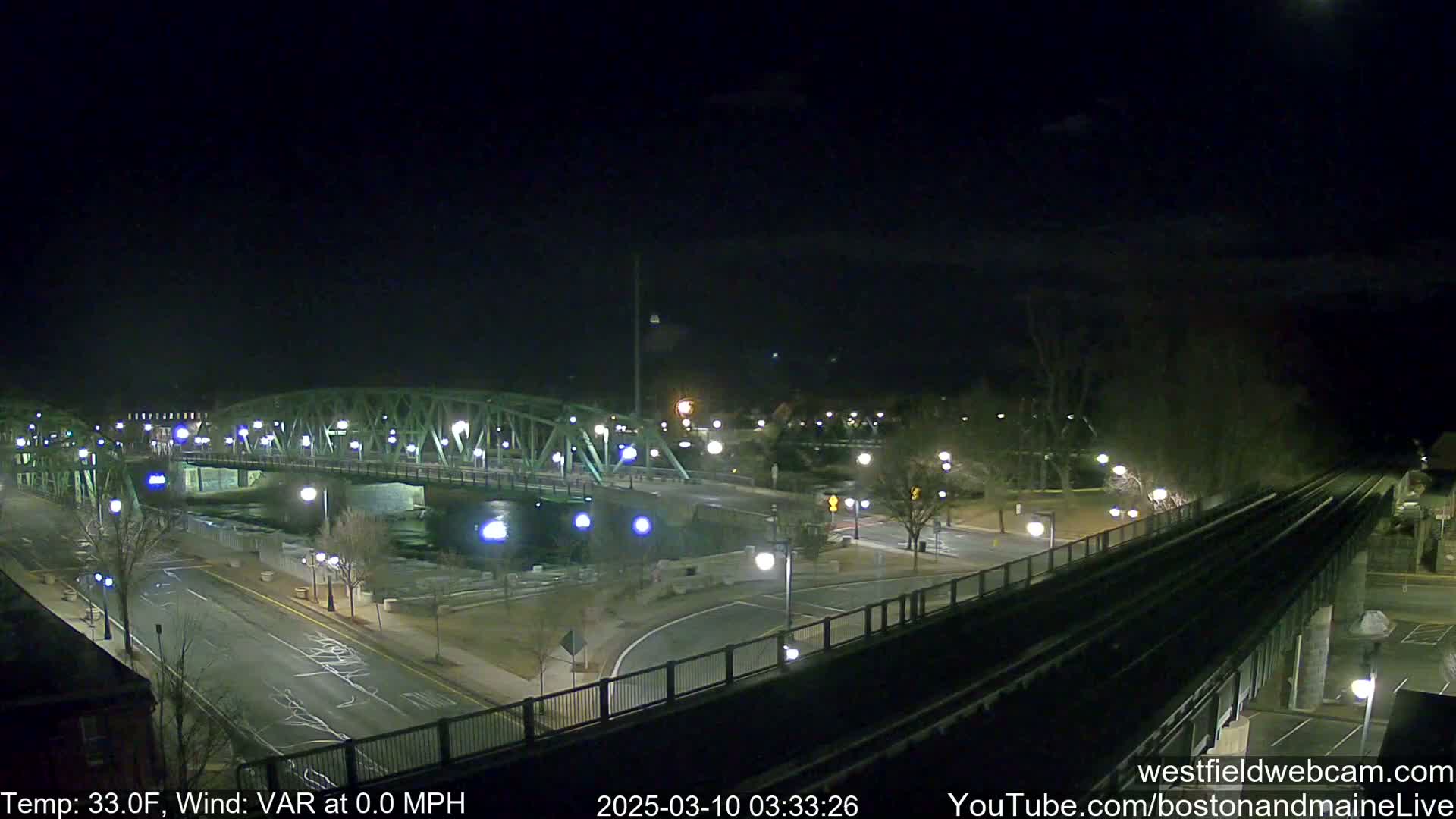 A nighttime view of a lit bridge over a dark river, with roadways and train tracks visible under a clear, dark sky.
