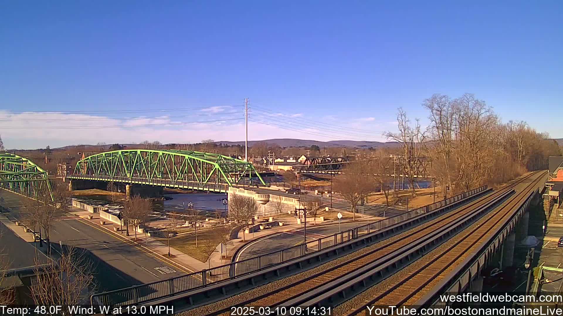 A bright green bridge spans a river, with train tracks and bare trees visible in the foreground under a clear blue sky.