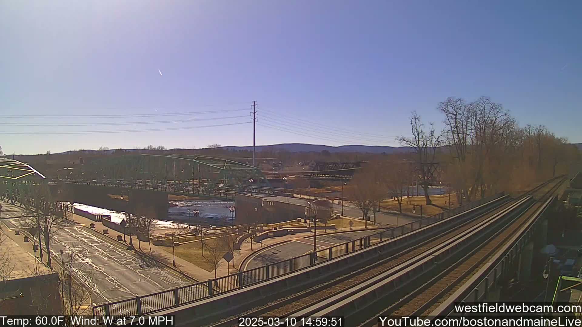 A green bridge spans a river, with train tracks and a paved road visible, under a clear, sunny sky with a 7 mph westerly wind and a temperature of 60°F.