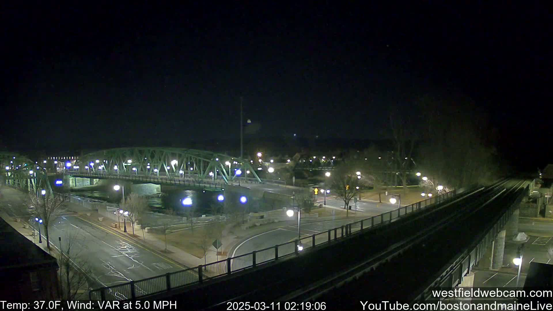 A nighttime view of a bridge over a river, with a road and train tracks visible, under a clear, dark sky.