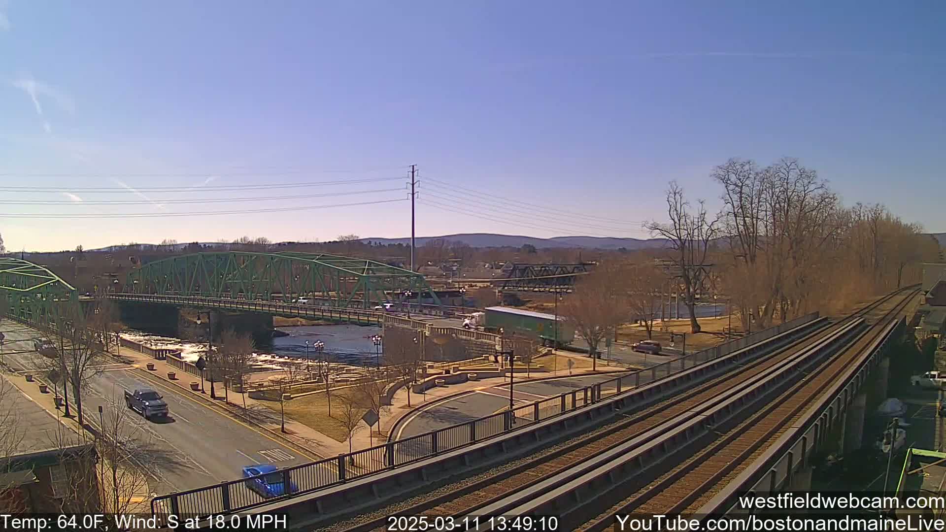 A high-angle view of a river with two bridges, one green and one dark-colored, alongside train tracks, under a clear, sunny sky.