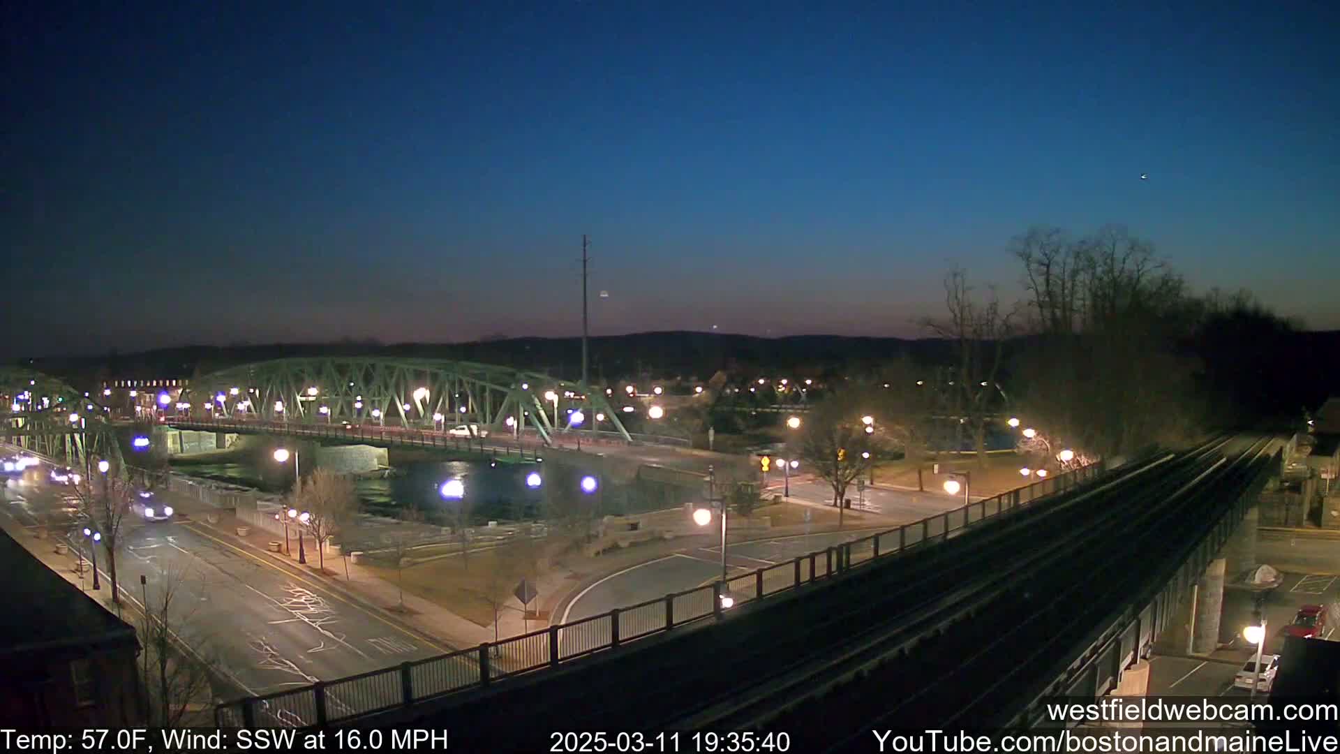 A nighttime view of a bridge over a river, with a road and train tracks visible, under a mostly clear sky with a south-southwest wind at 16 miles per hour.