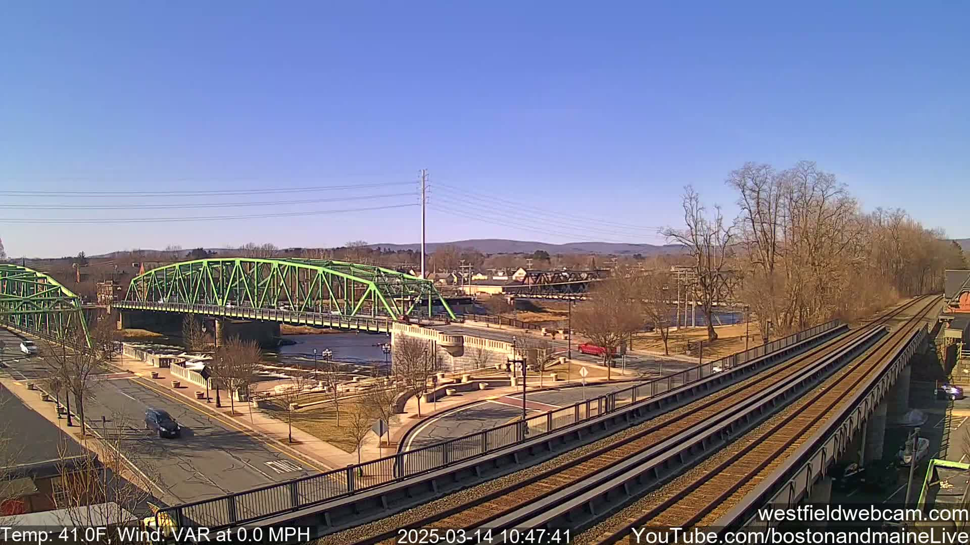 A sunny day shows two green bridges spanning a river, with train tracks and a road intersecting nearby, and bare trees lining the riverbank.