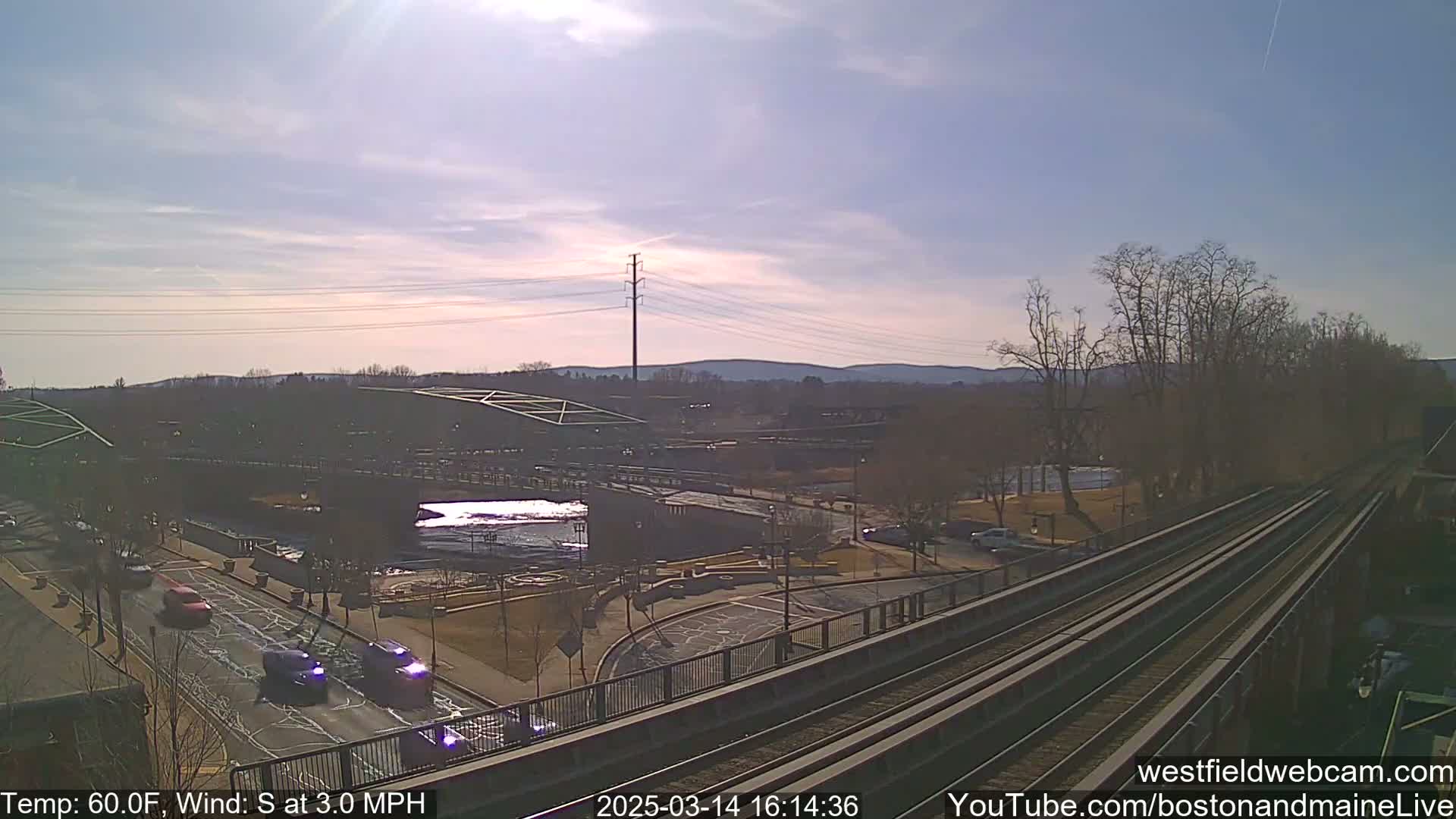 A sunny, slightly windy day is shown overlooking a bridge and a river, with train tracks and roadways visible below.