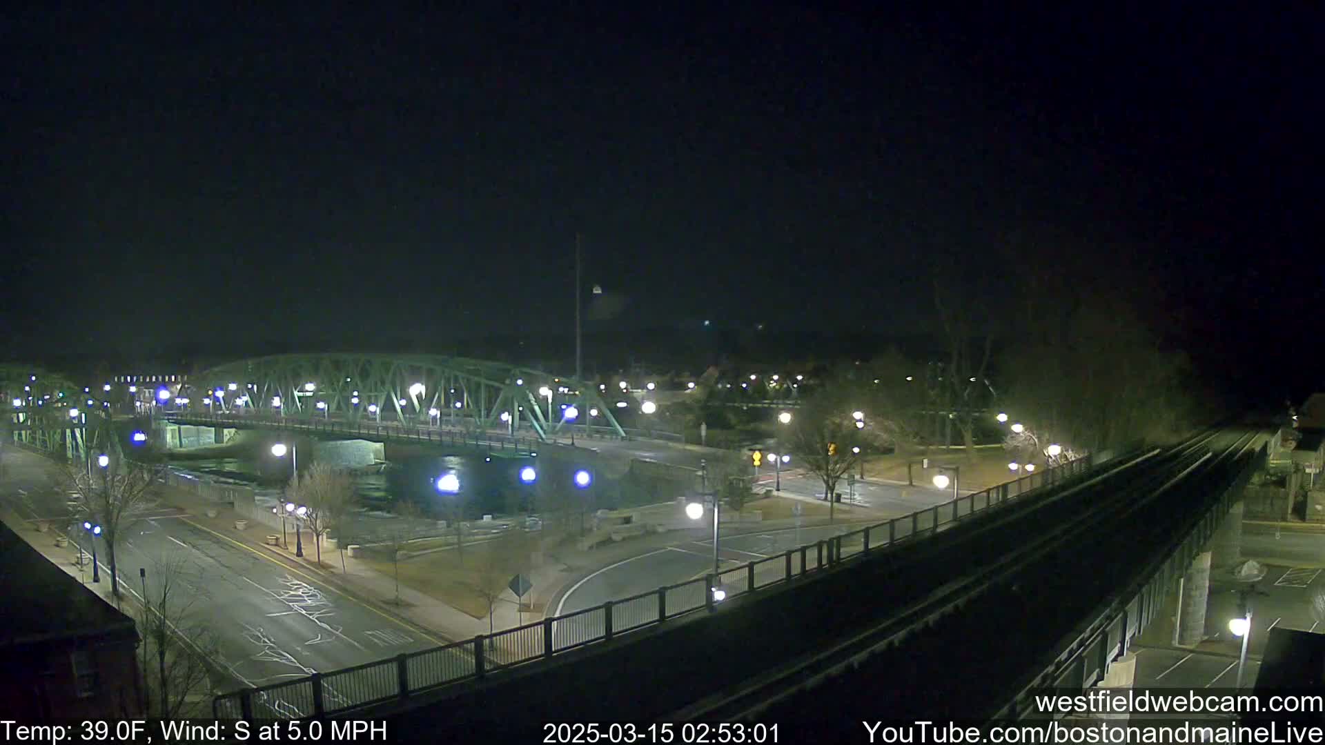 A nighttime view of a bridge over a river, with a road and train tracks visible, under a clear, dark sky with a temperature of 39°F and a south wind of 5 mph.